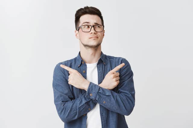 Waist-up shot of hesitant unsure attractive caucasian young man with bristle in glasses and blue shirt crossing hands on chest, pointing sideways as being doubtful what chose, looking right unsure.