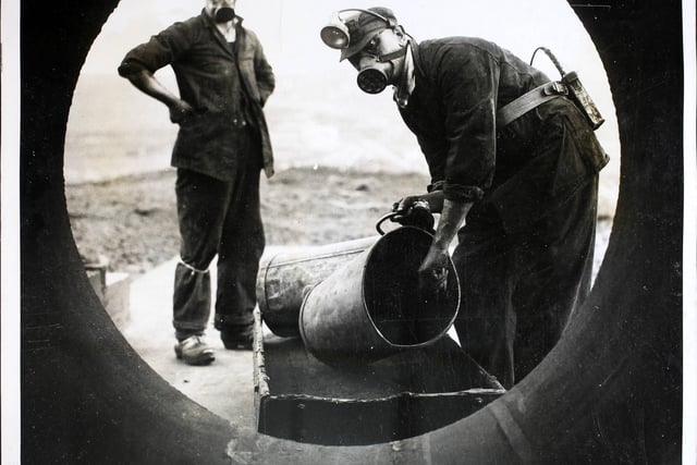Men wearing respirators loading a four-foot wide tube with coal dust on a hilltop at Harpur Hill, Buxton, before detonating it in a controlled explosion for an experiment. A handful of men at the site would be fighting continually against the causes of coal mine disasters. Coal dust, under certain conditions, is as dangerous as high explosive and these men were experimenting every day to find an effective method of combatting the menace.